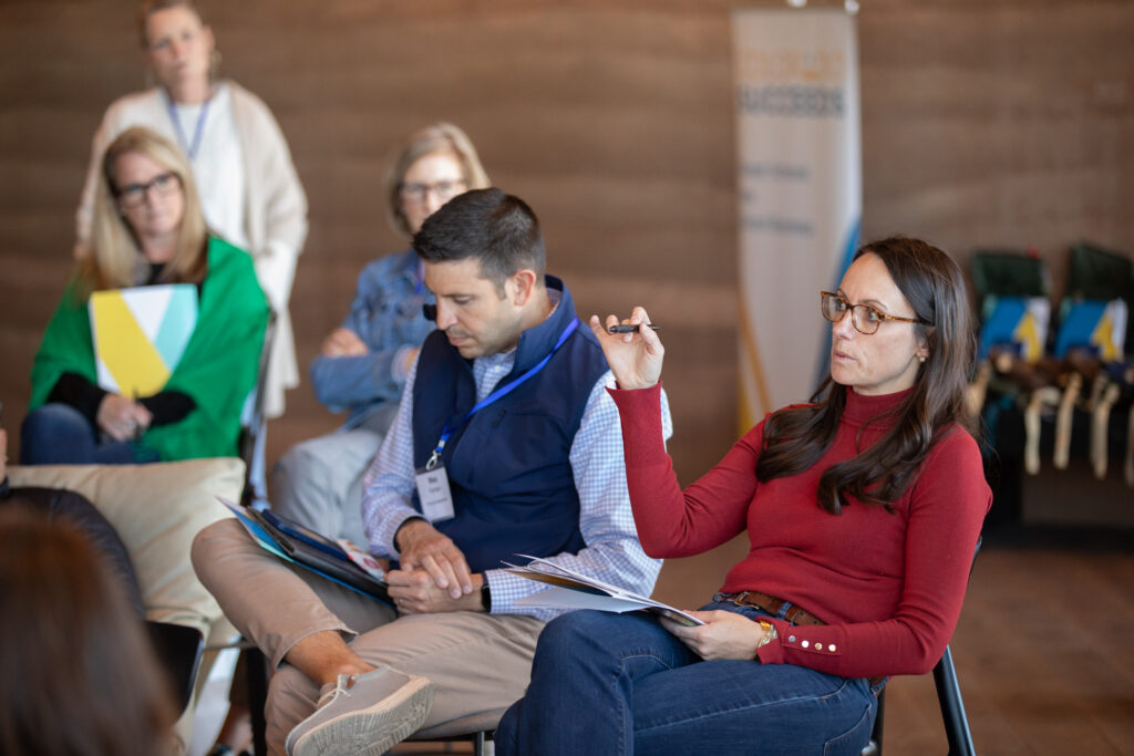 Colorado Succeeds team members participating in a group discussion at a retreat, with one Ashley raising a point while others take notes