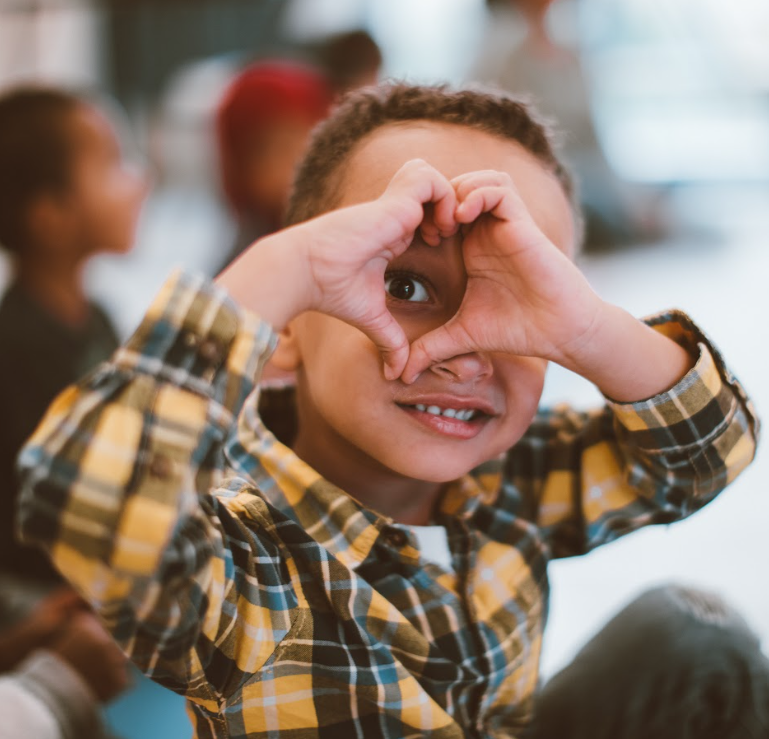 Young child smiling and making a heart shape with their hands in a classroom setting