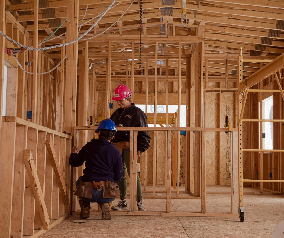 Two high school students wearing hard hats and tool belts working on wood framing inside a building under construction