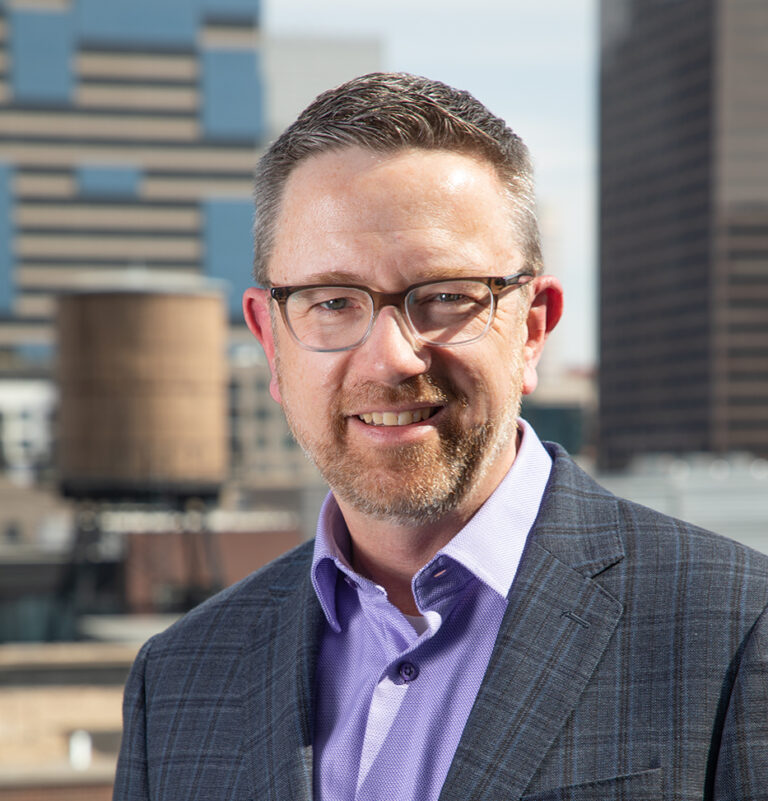Professional headshot of Joe Kuntner in a plaid jacket and lavender shirt, standing in front of a city skyline