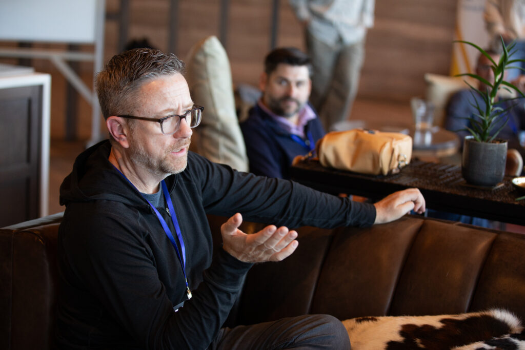 Joe Kuntner speaking while seated on a couch during a group discussion at a retreat, with other participants listening in the background