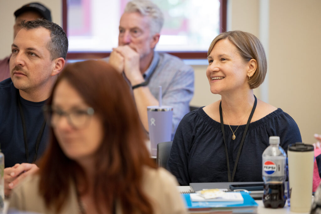 A woman smiles while participating in a professional learning session, surrounded by other engaged attendees with notebooks and drinks on the table