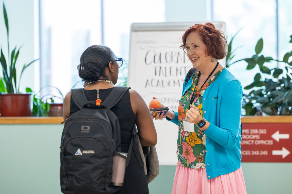 Two women talk in front of a whiteboard sign reading “Colorado Talent Academy.” One woman wears a bright blue cardigan and holds a peach while smiling. The other has a backpack and faces away. Potted plants and directional signage appear in the background.