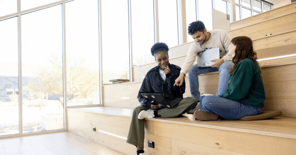 Three college students sit on wooden bleachers in a sunlit, modern campus building. One student works on a laptop while the others lean in, smiling and pointing at the screen, collaborating on a project.