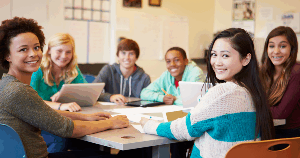 A diverse group of high school students sits around a table in a classroom, smiling toward the camera. Laptops, notebooks, and papers are spread out as they collaborate on a project together.