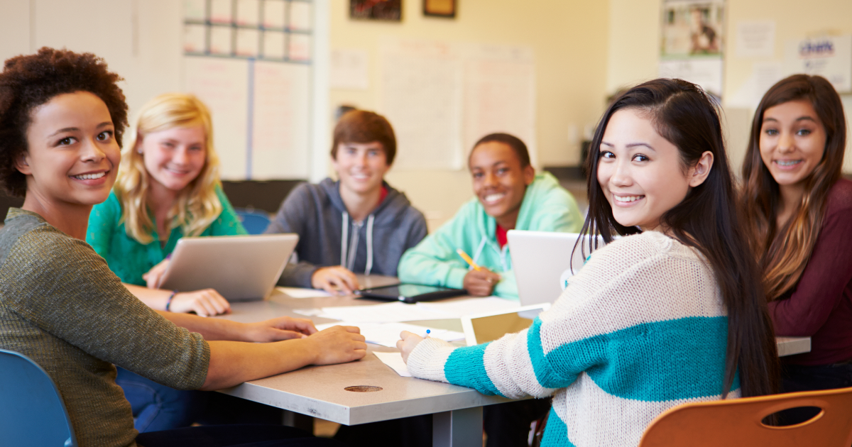 A diverse group of high school students sits around a table in a classroom, smiling toward the camera. Laptops, notebooks, and papers are spread out as they collaborate on a project together.