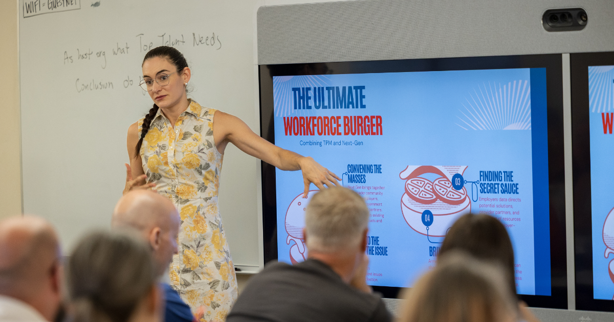 A woman presents to a seated audience in front of a screen titled 