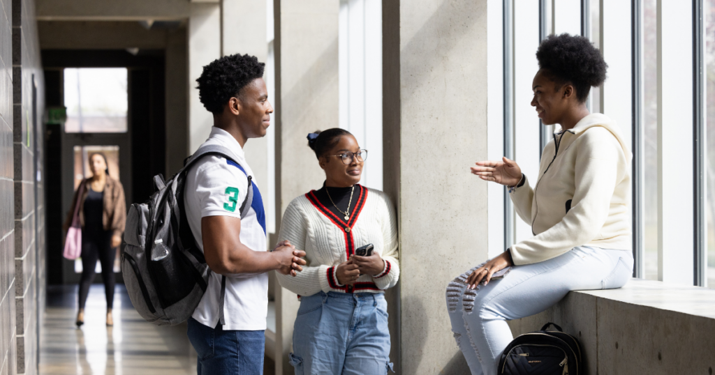 Three high school students talk and laugh together in a sunlit hallway. Two stand near a wall of tall windows while the third sits on a ledge, all appearing engaged and at ease. Backpacks and casual attire suggest a relaxed school setting.