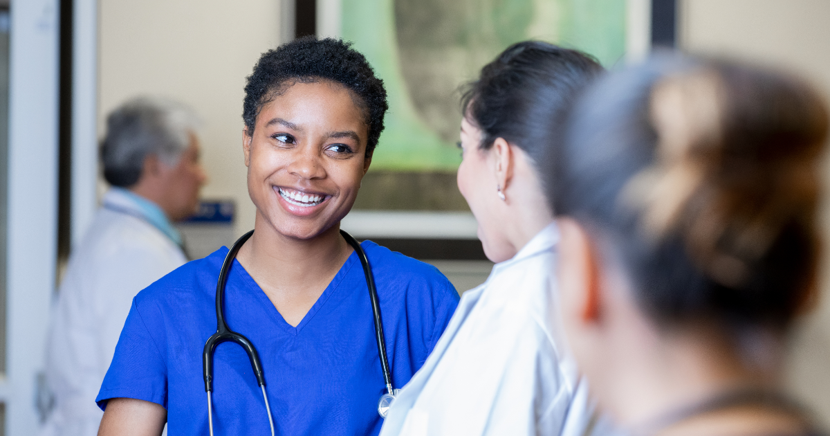female nurse in blue scrubs smiling at a female doctor in a white coat with her back to the camera